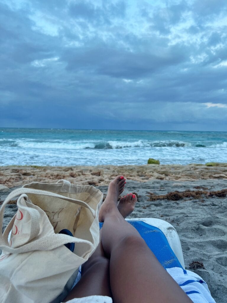 Relaxing beach scene with a close-up of a person's legs resting on a blue surfboard, toes painted red, as they recline on a sandy beach. A canvas tote bag lies beside them, and the ocean waves meet a cloudy sky in the background, creating a peaceful seaside setting.
