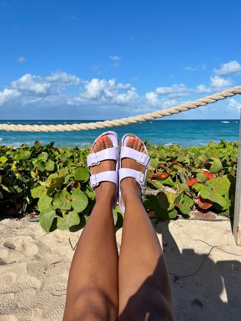 View of a person's legs extended in front, wearing purple sandals with the backdrop of a sandy beach, lush greenery, and a bright blue sky. The person is seated on a beach with a thick rope barrier leading to the ocean in the foreground