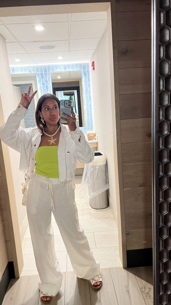 A woman takes a mirror selfie in a stylish restroom. She's dressed in a white jacket and trousers paired with a bright lime green top. Accessories include a starfish necklace, large hoop earrings, and a chic shoulder bag. She poses with a peace sign, adding a playful vibe to her trendy outfit.