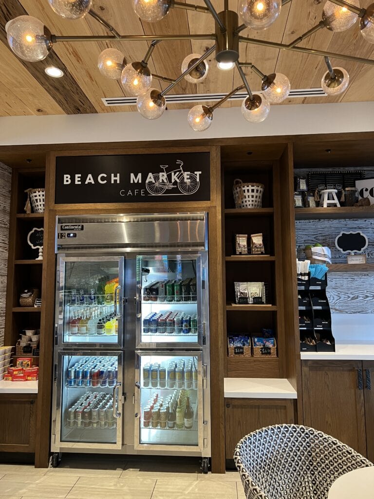 View inside the Beach Market Cafe showing a well-stocked refrigerator with assorted beverages and snacks, flanked by shelves with various goods, under a wooden ceiling illuminated by a modern chandelier.