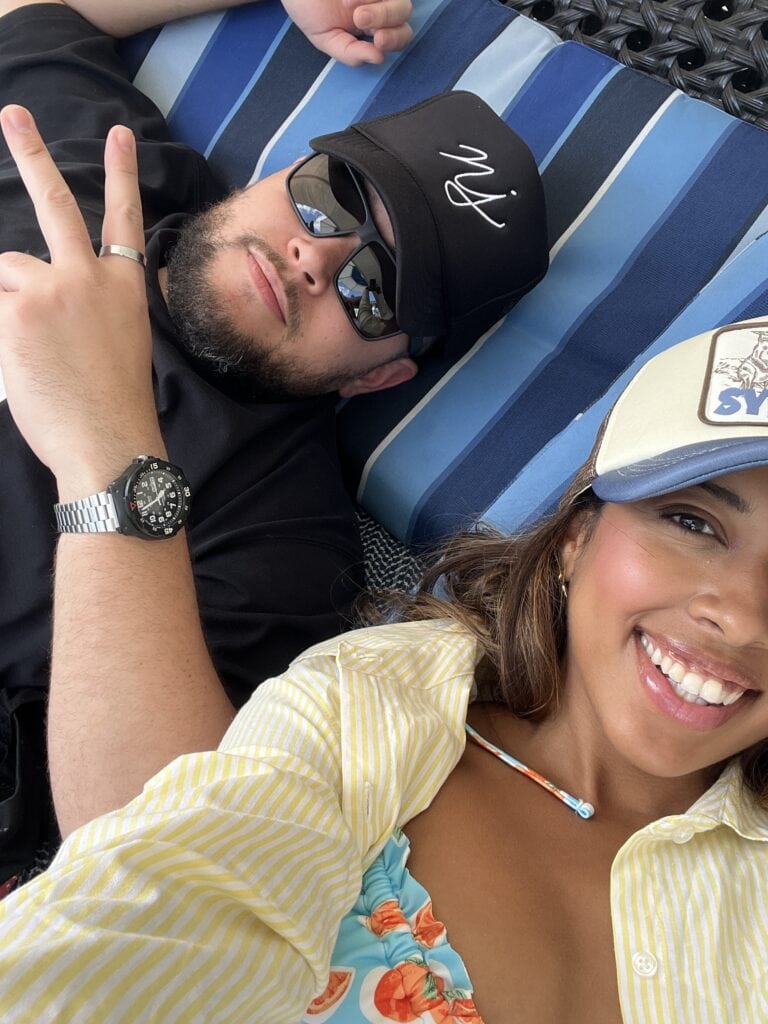 A cheerful selfie of a couple relaxing outdoors on a blue-striped lounger. The man, wearing sunglasses and a black cap, flashes a peace sign, while the woman, smiling brightly, sports a yellow-striped shirt over a floral bikini top. The scene captures a casual, happy moment between the pair.